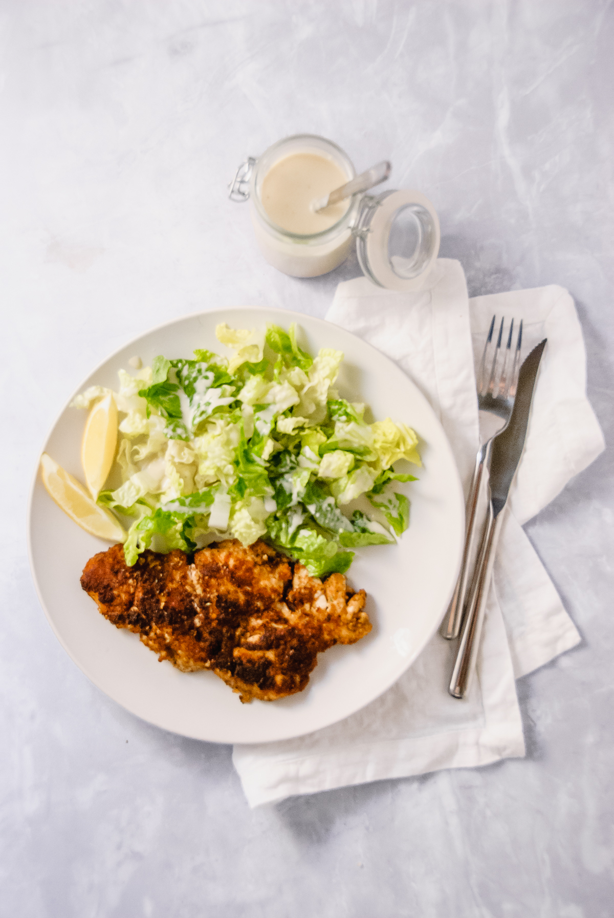 White plate with a caesar salad: romaine lettuce and light caesar salad dressing, lemon slices and chicken cutlets: breaded chicken fried in garlic infused oil.